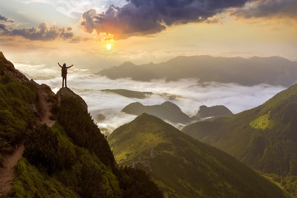 Wide mountain panorama. Small silhouette of tourist with backpack on rocky mountain slope with raised hands over valley covered with white puffy clouds. Beauty of nature, tourism and traveling concept